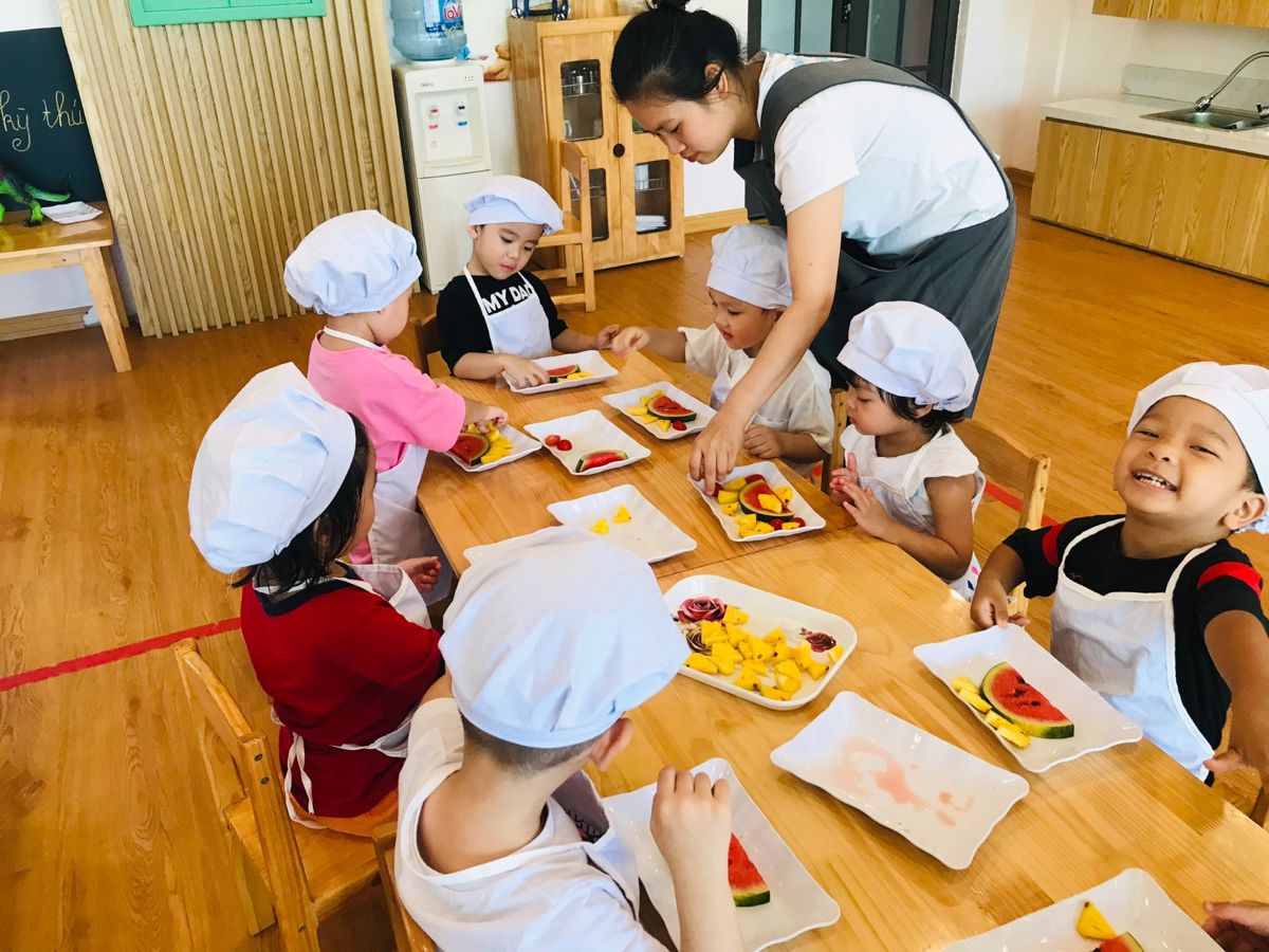 Các bé đội mũ đầu bếp bày dưa hấu và dứa ra đĩa cùng cô